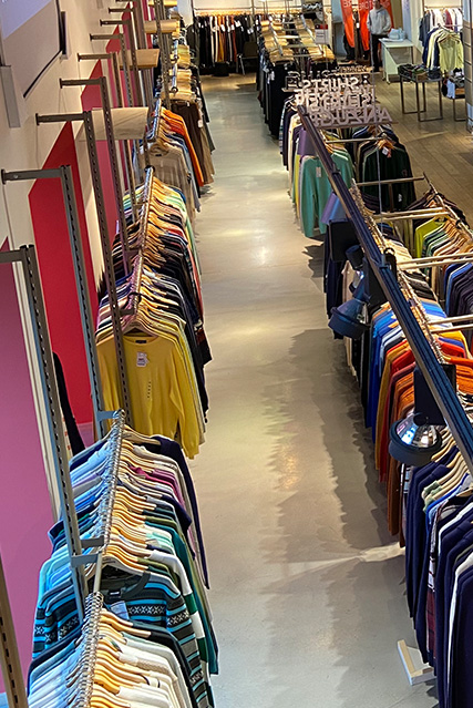Ein langer schmaler Gang in einem Bekleidungsgeschäft mit bunten Kleidungsstücken, die ordentlich auf beiden Seiten aufgereiht sind.