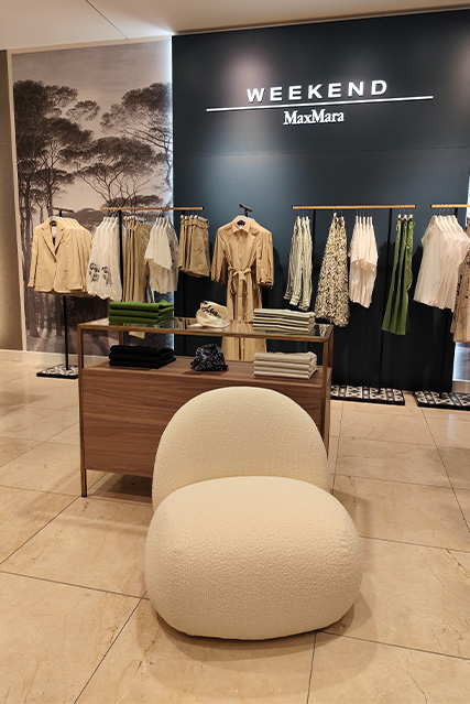 Ein eleganter Modestore mit dem Schriftzug "WEEKEND MaxMara" an der Wand, zeigt eine Auswahl von Kleidung in neutralen Tönen, aufgehängt an Kleiderstangen, mit einem cremefarbenen Sessel im Vordergrund.