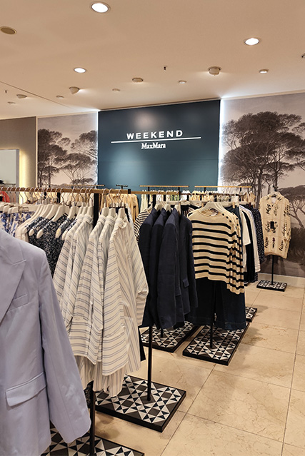 Kleidungsausstellung in einem Geschäft, mit gestreiften und einfarbigen Oberteilen auf Kleiderständern; im Hintergrund ein Schild mit der Aufschrift "Weekend MaxMara".