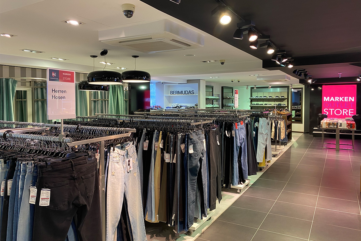 Das P&C Markenoutlet in Bremen mit der Abteilung für Herrenhosen, auf Kleiderständern präsentiert; im Hintergrund weitere Kleidung und ein Schild mit der Aufschrift "Marken Outlet Store".