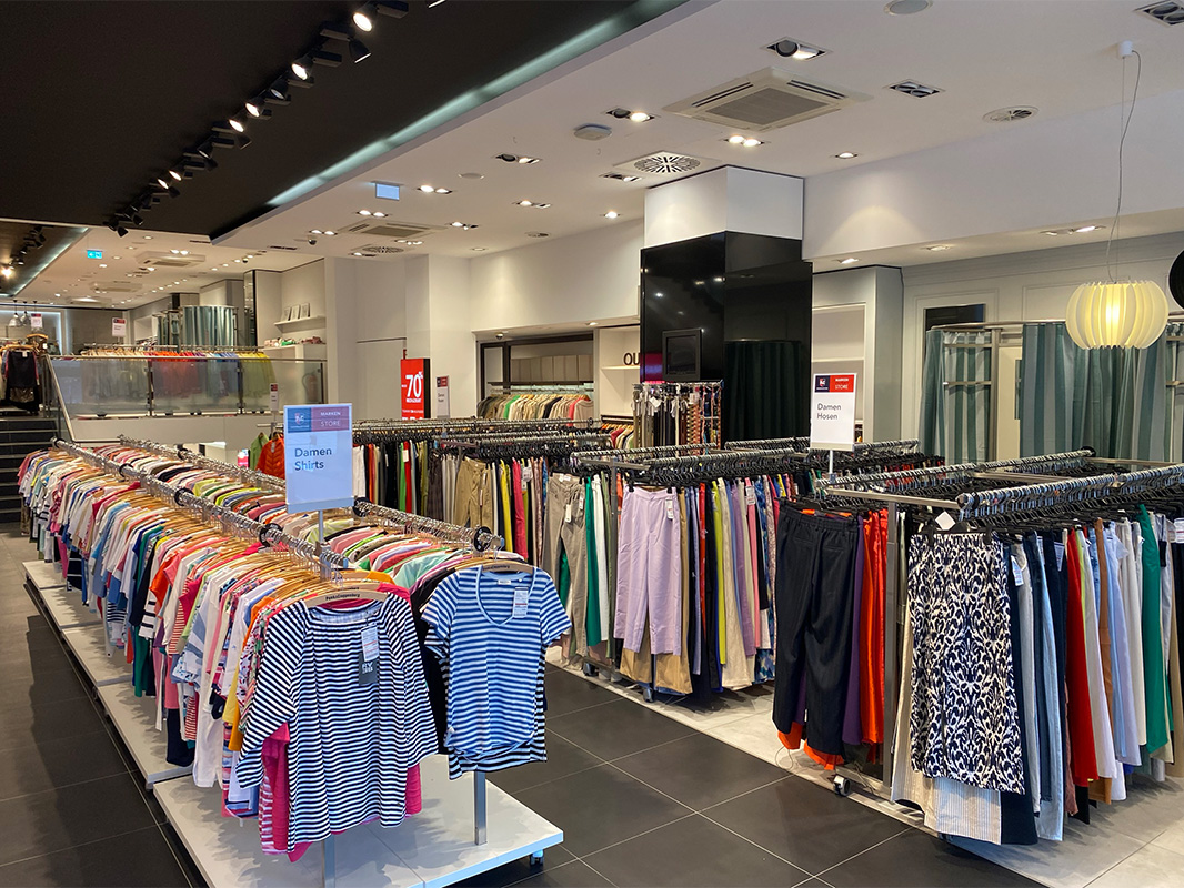 Das P&C Markenoutlet in Bremen mit Reihen von Damenoberteilen und -hosen auf Kleiderständern, einem Schild mit der Aufschrift „Damen Shirts” und einem Sale Schild im Hintergrund.