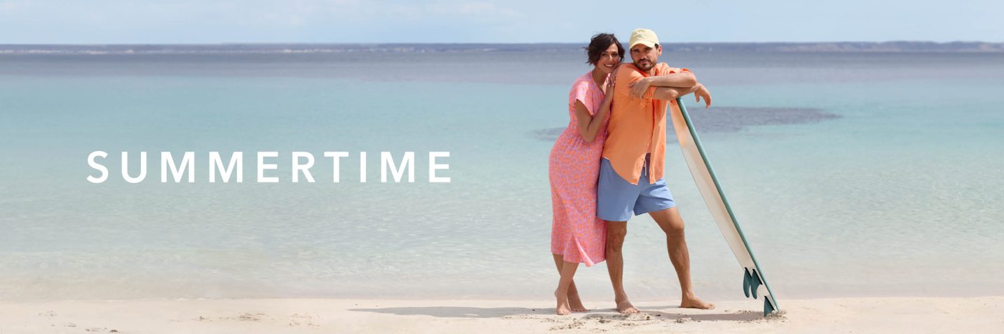 Ein Paar steht am Strand unter dem Schriftzug "SUMMERTIME" neben einem Surfbrett mit Blick auf das Meer.